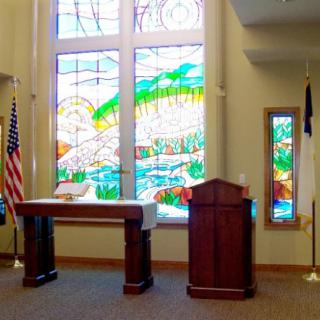 A chapel interior featuring stained glass windows, a podium, an altar, and flags.