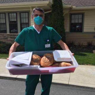 A medical professional wearing scrubs and a face mask holds a tray of freshly baked bread outside a healthcare facility.
