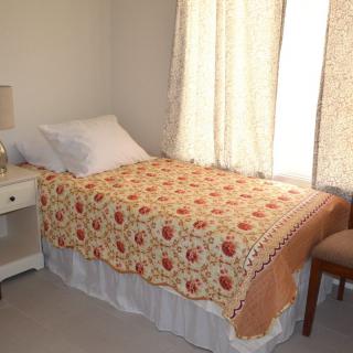 A small bedroom with a single bed covered by a patterned quilt, next to a lamp and chair.