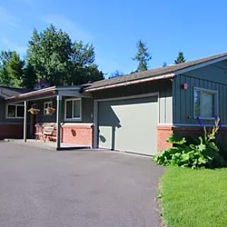 A single-story house with a garage and driveway surrounded by green lawn and trees under a clear blue sky.