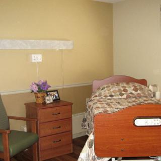 A hospital room with a bed, a side table with flowers and a picture frame, and a chair.