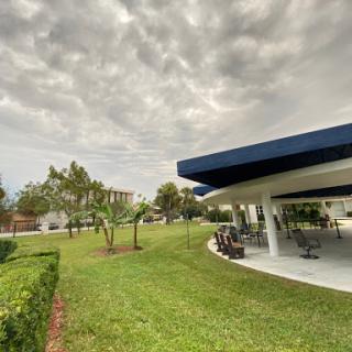 A scenic view of a park featuring lush green grass, trees, and a covered outdoor seating area under a cloudy sky.
