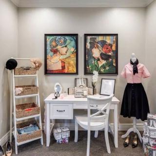 A stylish vanity area with vintage decor, featuring a makeup desk, a mannequin with retro clothing, and colorful paintings.