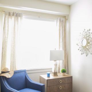 A cozy corner with a blue armchair, a side table with a lamp, and light curtains framing a large window.