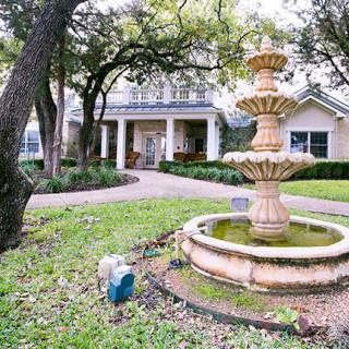 A scenic view of a house with a three-tiered fountain in the front yard, surrounded by trees and greenery.