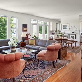 A spacious and well-lit living room featuring comfortable seating, a patterned rug, and large windows overlooking greenery.