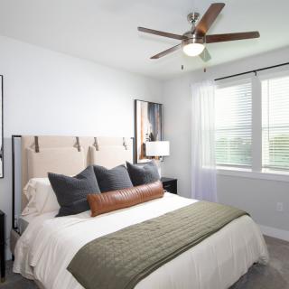 A modern bedroom with neutral tones, featuring a stylish bed, ceiling fan, and ample natural light.