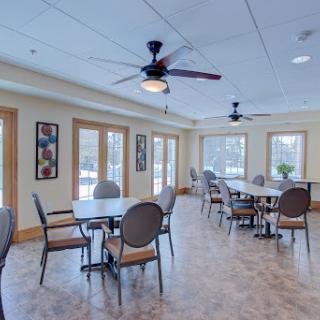 A spacious community dining area featuring tables, chairs, and ceiling fans, illuminated by natural light from large windows.