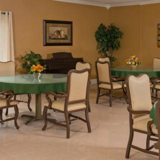 A welcoming dining room setup featuring several green-topped tables surrounded by beige chairs, enhanced by decorative plants and a floral centerpiece.