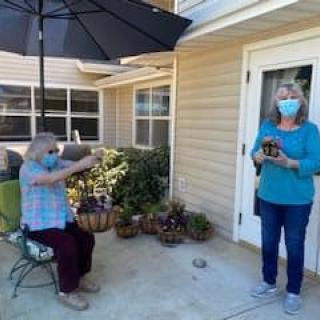 Two people practicing social distancing while wearing masks, one sitting and the other standing in a garden setting.