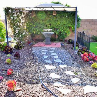 A garden with a stone pathway leading to a pergola and fountain, surrounded by gravel and plants.