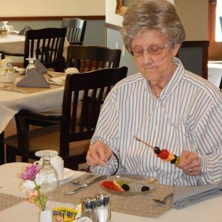 An elderly woman prepares a fruit skewer at the dining table.