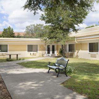 A serene outdoor area featuring a building, pathways, and a bench surrounded by greenery.