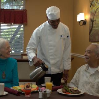 A chef serving two seniors at a dining table.