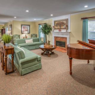 A spacious living room featuring a grand piano, cozy sofas, and a fireplace, elegantly decorated with artwork and greenery.