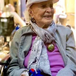An elderly woman sitting in a wheelchair, smiling and wearing a hat and scarf outdoors.