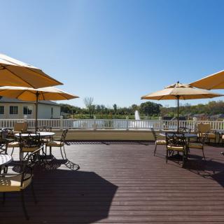 A sunny outdoor deck area with tables, chairs, and umbrellas overlooking a lake.