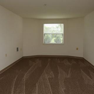 An empty room with a single window and carpeted flooring, illuminated by natural light.