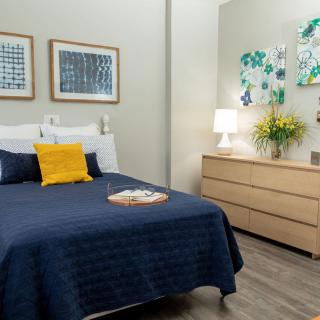 A well-decorated bedroom featuring a bed with blue bedding and a yellow pillow, framed artwork on the walls, and a wooden dresser with decorative items.