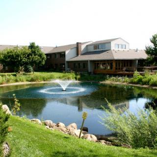A serene landscape featuring a tranquil pond with a fountain, surrounded by lush greenery and a modern building.