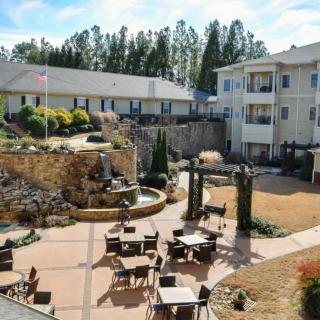 A sunny outdoor courtyard featuring a seating area with tables and chairs, surrounded by landscaped gardens and adjacent buildings.