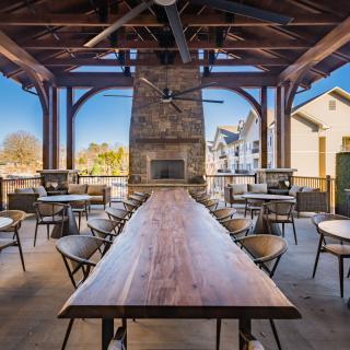 An inviting outdoor dining area featuring a long wooden table, surrounded by chairs, under a covered patio with a stone fireplace.