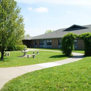 A tranquil outdoor area with a pathway leading to a school building, surrounded by greenery and seating benches.