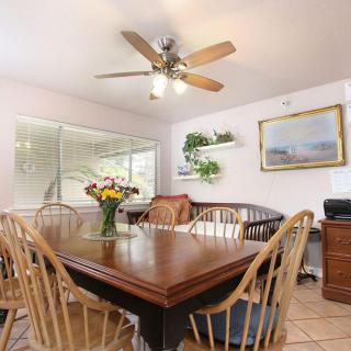 A cozy dining room with a wooden table, surrounded by chairs and topped with a bouquet of flowers. A ceiling fan hangs above, and there's ample natural light from the window.