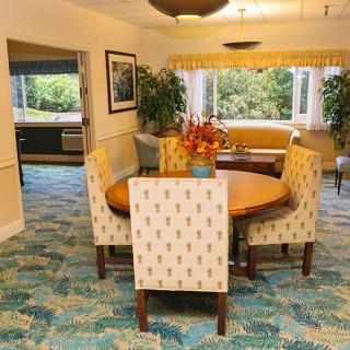 A cozy dining room with a round table and upholstered chairs, featuring a patterned carpet and large windows allowing natural light.