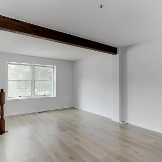 An empty room with wooden flooring and a large window, featuring exposed beams and white walls.