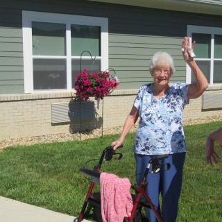 An elderly woman waves cheerfully while standing outside with her walker on a sunny day.