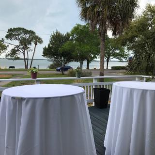 A scenic outdoor view with tables covered in white tablecloths, set against a backdrop of trees and waterfront.