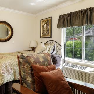A cozy bedroom featuring a bed with decorative pillows, an armchair, and a large window allowing natural light to fill the room.