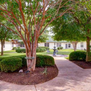 A serene garden with a pathway lined by trees and bushes, leading to a fountain in the center.