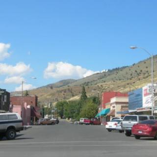A small town street lined with parked cars and shops, set against a mountainous background under a clear blue sky.
