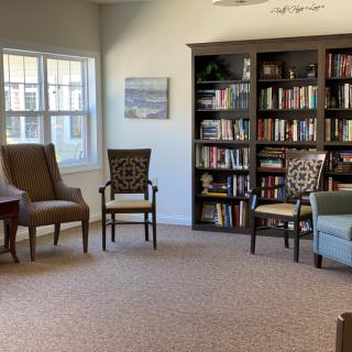 A cozy reading room with bookshelves, various chairs, and natural light from a window.