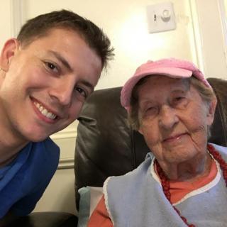A young man takes a selfie with an elderly woman indoors, both smiling.