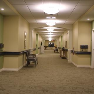 A well-lit hallway with carpeted flooring and chairs along the sides.