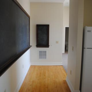 A narrow hallway features a large chalkboard on one wall, with a refrigerator and hardwood flooring.