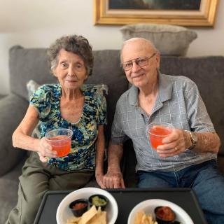 An elderly couple sitting on a sofa, enjoying drinks and snacks.