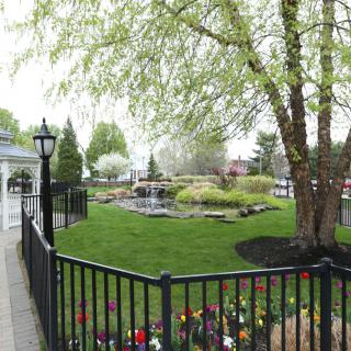 A picturesque garden scene with a black fence, lush green lawn, colorful flowers, and large trees.