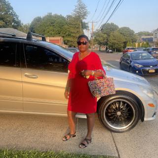 A stylish woman in a red dress poses beside her silver car, enjoying a sunny day.