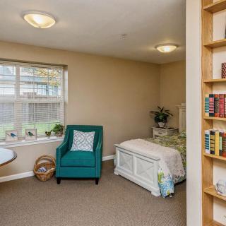 A cozy bedroom with a teal armchair, a single bed, and a bookshelf filled with books, illuminated by natural light.
