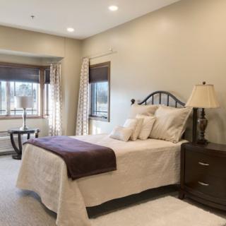 A cozy bedroom with a neatly made bed, bedside lamp, and large windows with curtains.