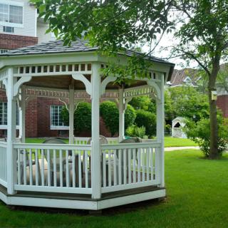 A white gazebo standing on a lush green lawn surrounded by trees and vegetation in a garden setting.
