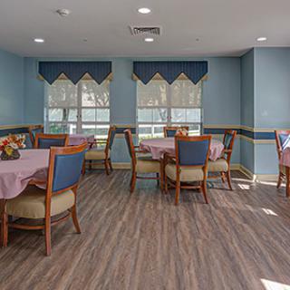 A cozy dining room interior with neatly arranged tables and chairs, featuring elegant curtains and a wooden floor.