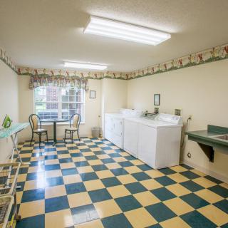 A well-lit laundry room with checkered flooring, featuring washing machines and a small seating area by the window.