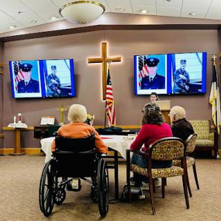 A gathering of senior citizens in a community center paying tribute to veterans, featuring images displayed on television screens.
