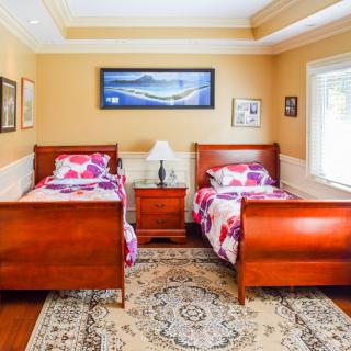 A cozy bedroom featuring two wooden twin beds with floral bedding and a central nightstand, adorned with paintings and a patterned carpet.