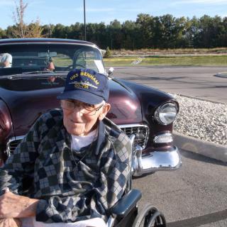 An elderly man in a wheelchair sits in front of a vintage car on a sunny day.